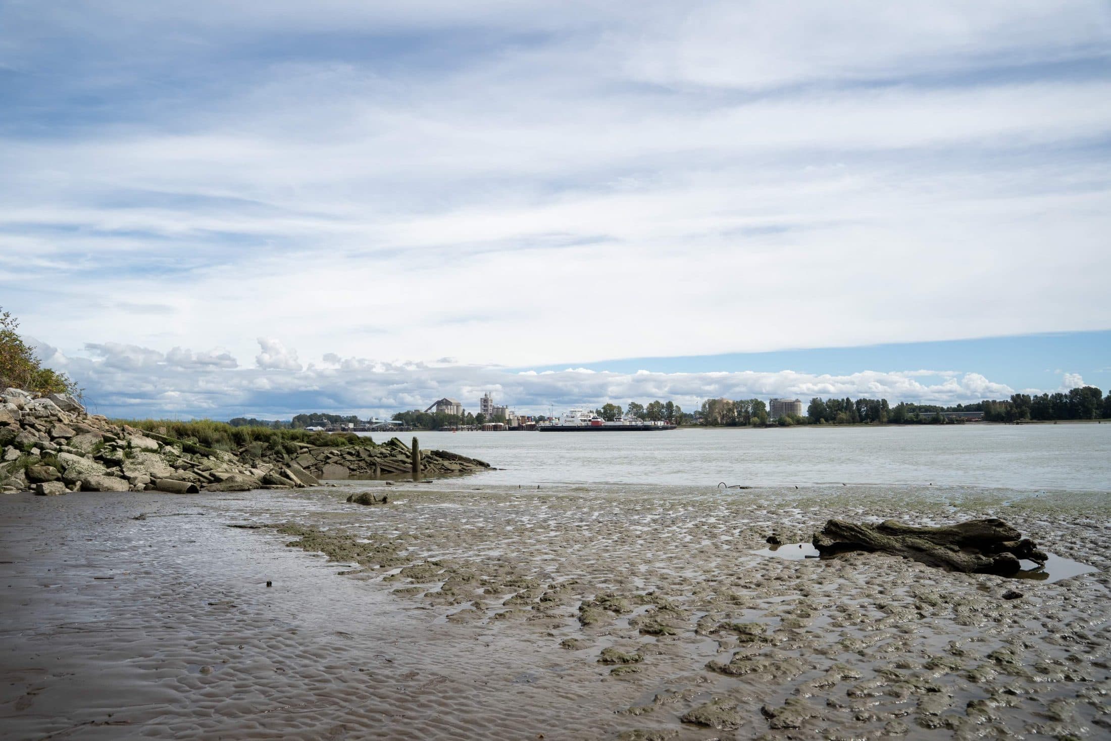 A rocky beach shore along a riverbank that looks out to buildings and clouds and sky