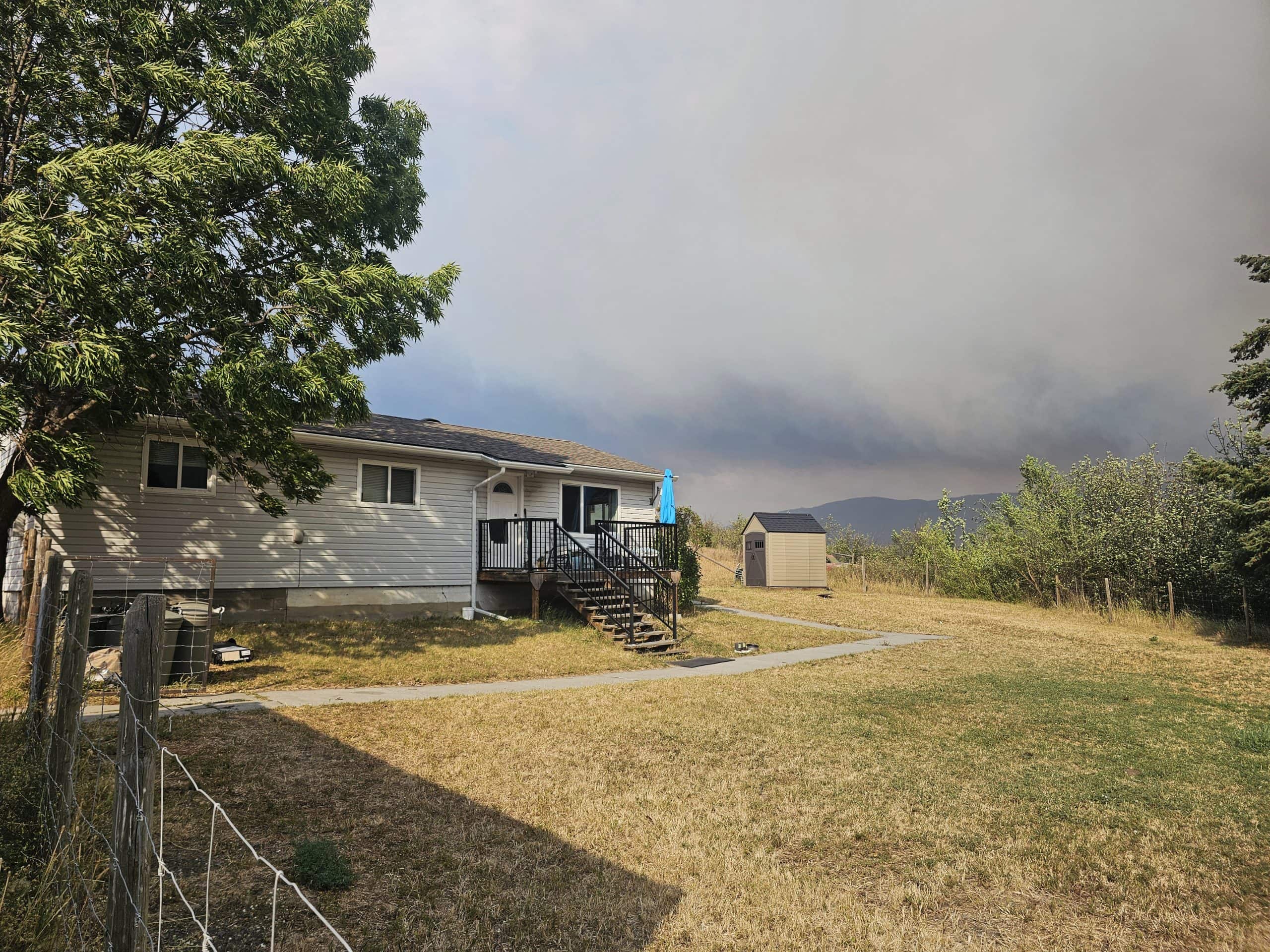 A grey house with a white door under a smoky, cloudy sky. In the background is a small shed and trees. The grass is dry.
