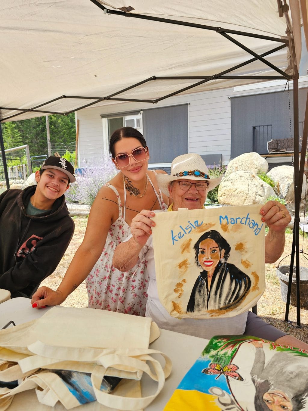 A child in a black hoodie, a woman in sunglasses and a sundress and an older woman in a white hat smile at the camera. The older woman holds up a painting of the younger woman. All three are under a white canopy.