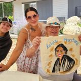 A child in a black hoodie, a woman in sunglasses and a sundress and an older woman in a white hat smile at the camera. The older woman holds up a painting of the younger woman. All three are under a white canopy.