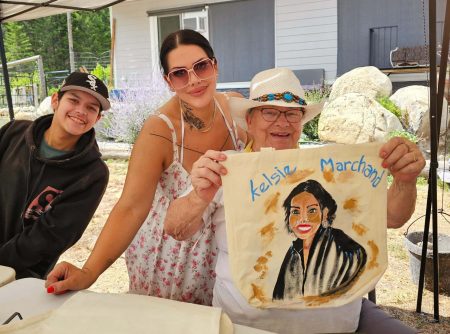 A child in a black hoodie, a woman in sunglasses and a sundress and an older woman in a white hat smile at the camera. The older woman holds up a painting of the younger woman. All three are under a white canopy.