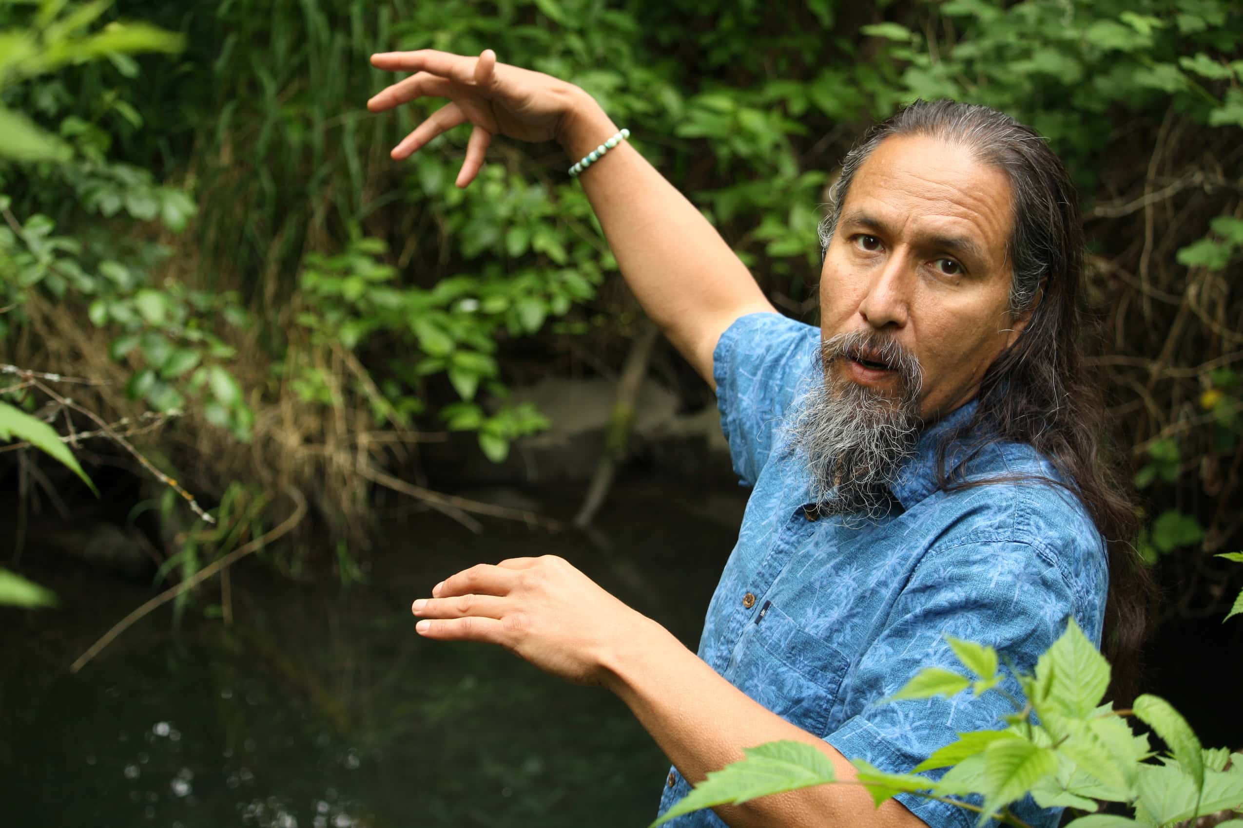 A man gesturing over a river with forest greenery surrounding it.