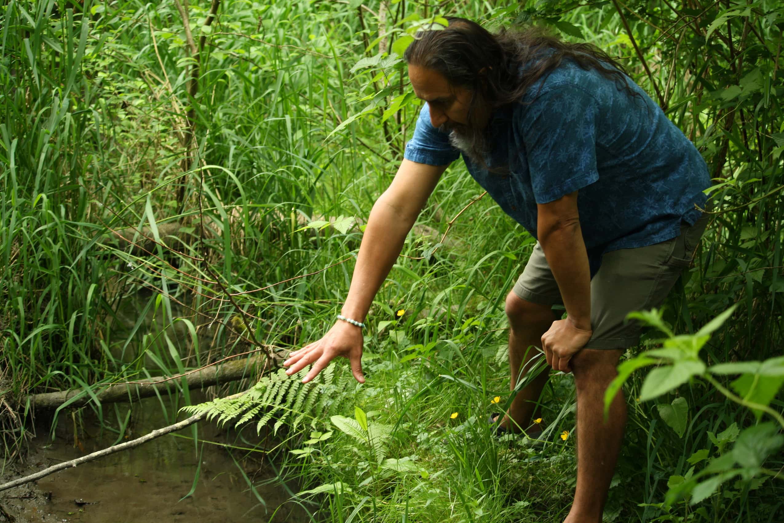 Tim Kulchyski leans over the edge of a riverbank with greenery behind him.
