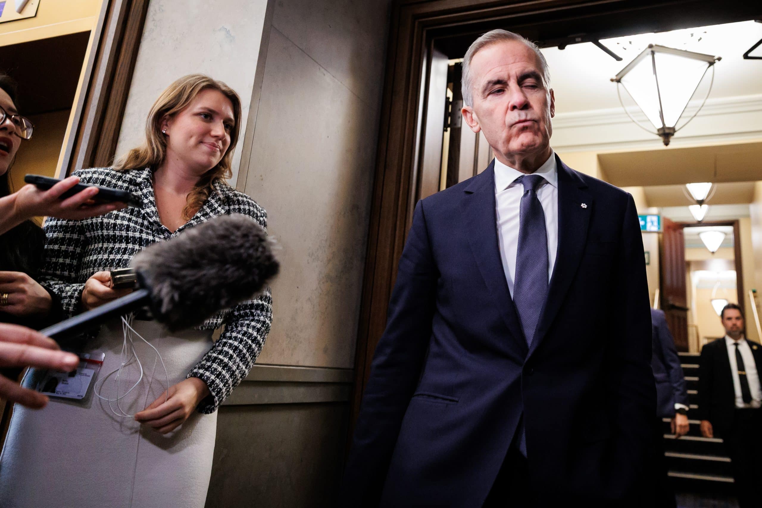 A man (Prime Minister Mark Carney) closes his eyes listening to questions from reporters with microphones in the middle of a doorway.