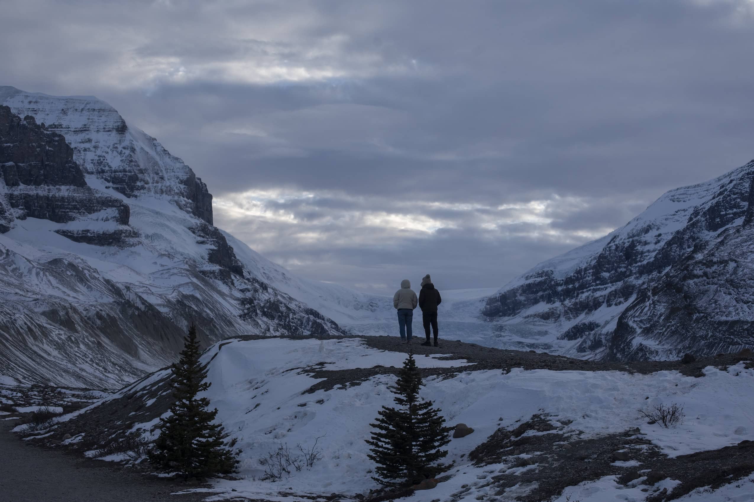 Seen from behind, two people stand and look out over a landscape of snowy mountains and a glacier.