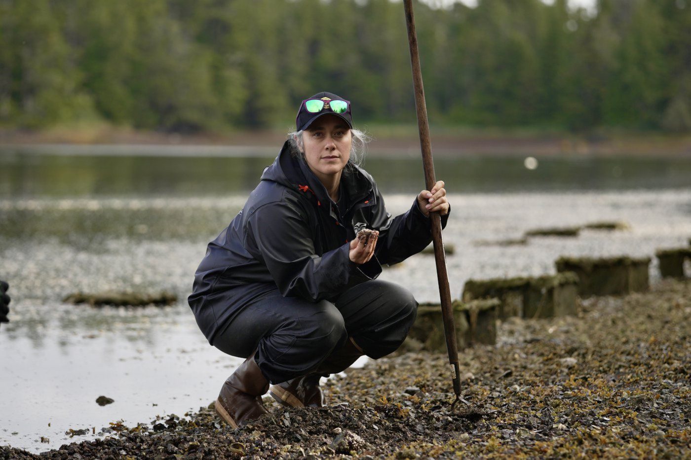 A woman crouches along a riverbank, holding a green crab in hand.