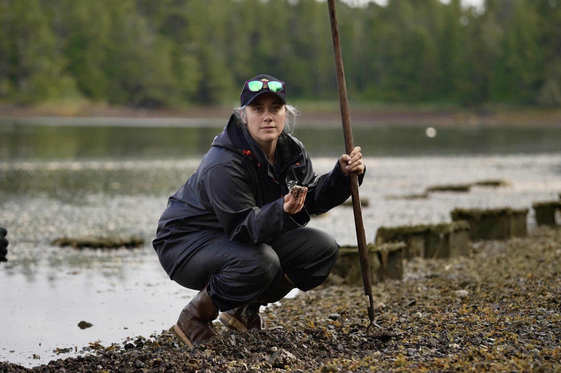 A woman crouches along a riverbank, holding a green crab in hand.