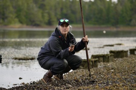 A woman crouches along a riverbank, holding a green crab in hand.