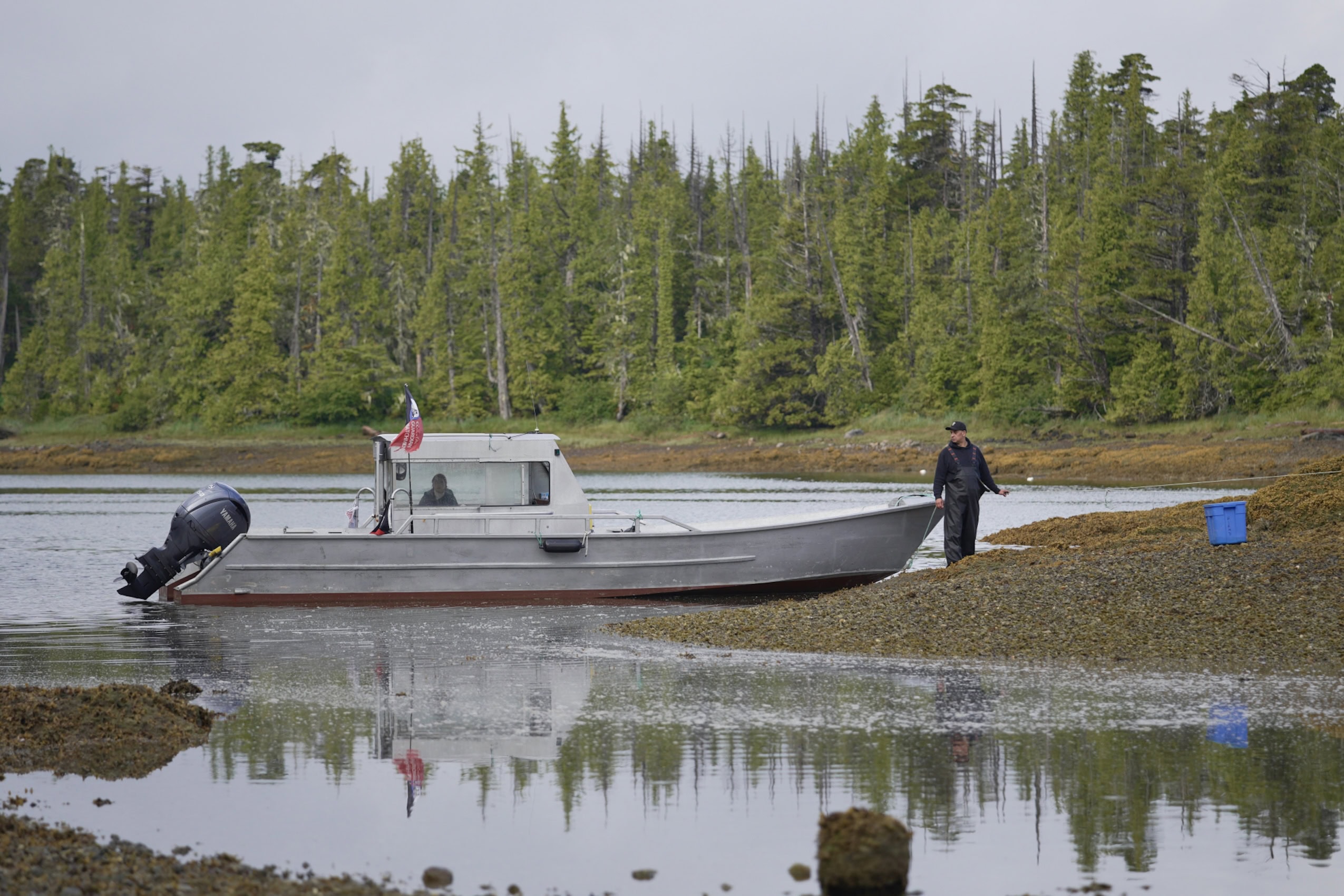 Small fishing boat docked on a rocky shoreline. A man in fishing overalls and a black baseball cap looks over his shoulder at the boat and the water.