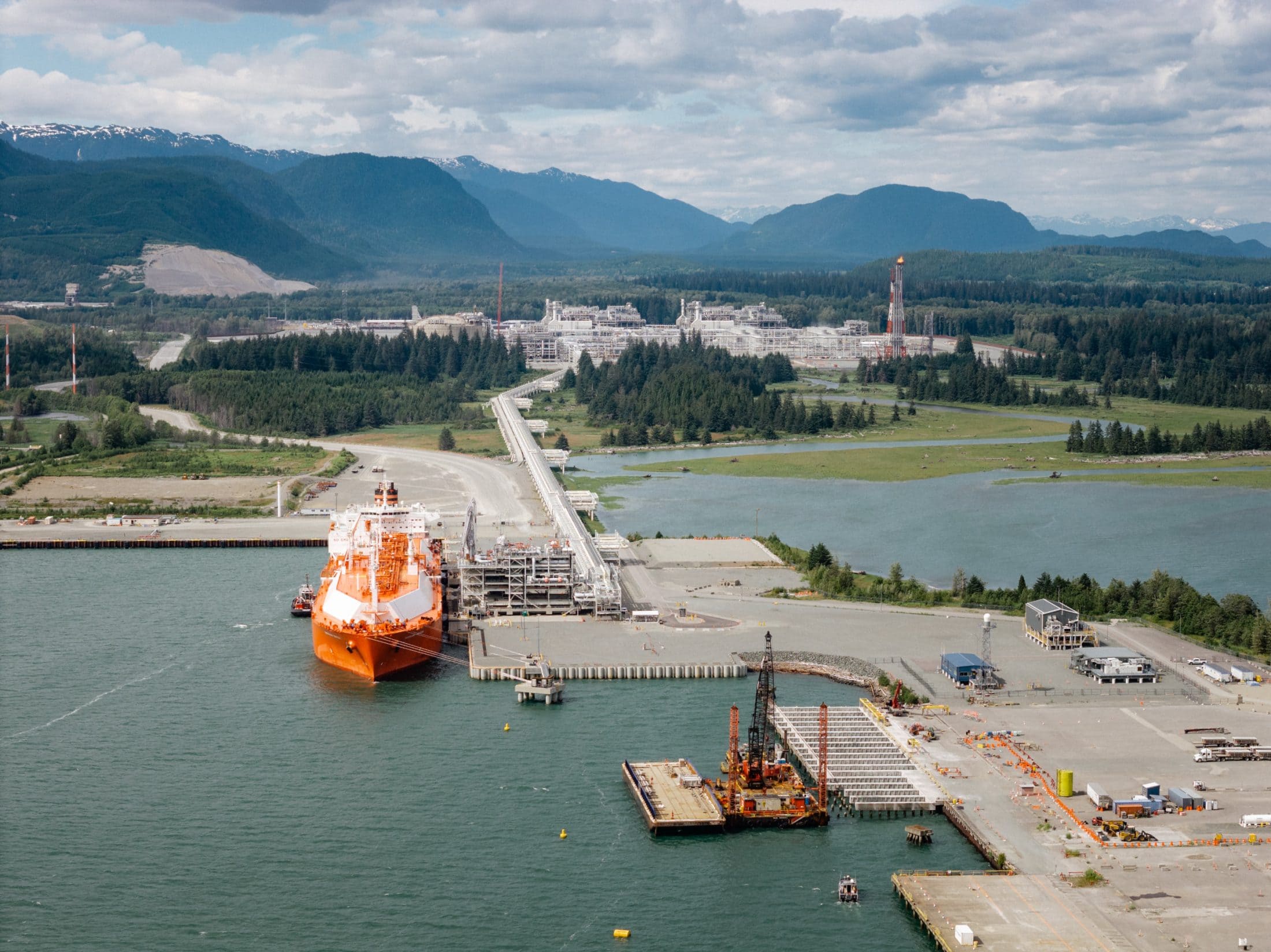 An orange boat sits in an industrial harbour with an LNG plant surrounded by trees and mountains in the background