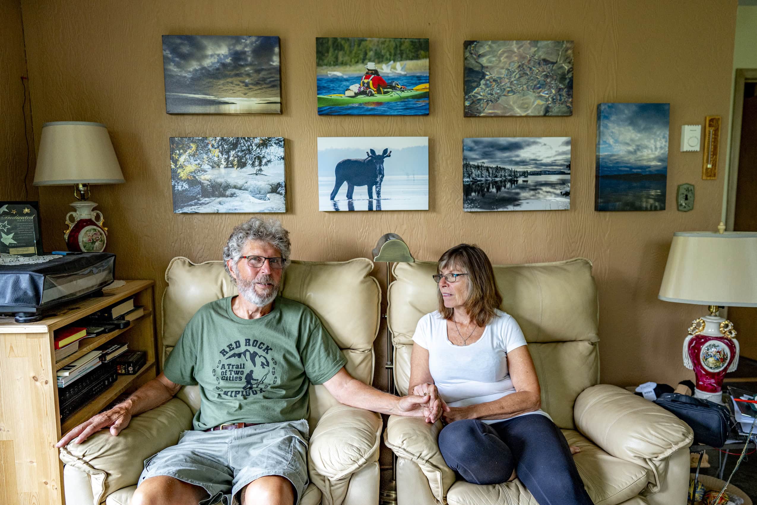 A couple sits on a beige couch, in front of a wall of photographs