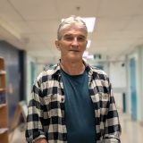 A man wearing a plaid shirt stands in a hallway looking at the camera