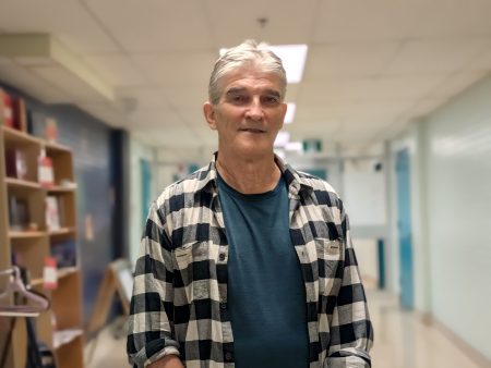 A man wearing a plaid shirt stands in a hallway looking at the camera