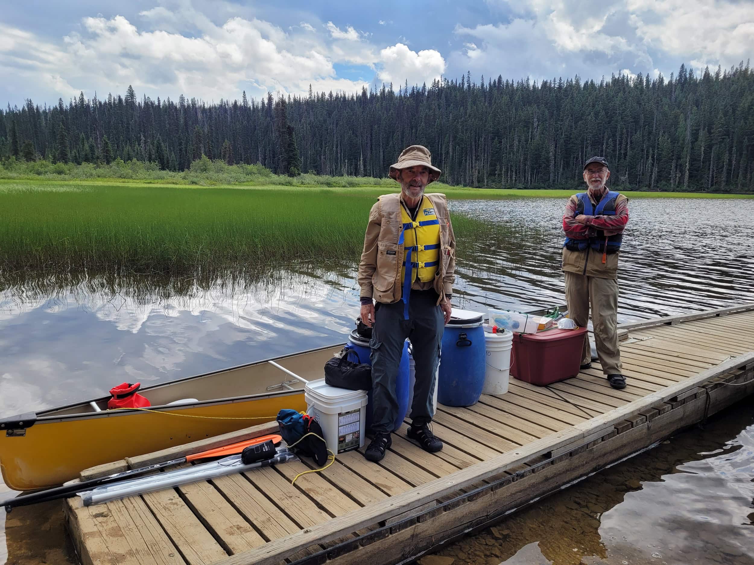 Two older men are seen posing on a dock, wearing hats and lifejackets with a canoe and gear. A marshy background with trees and sun and clouds is behind.
