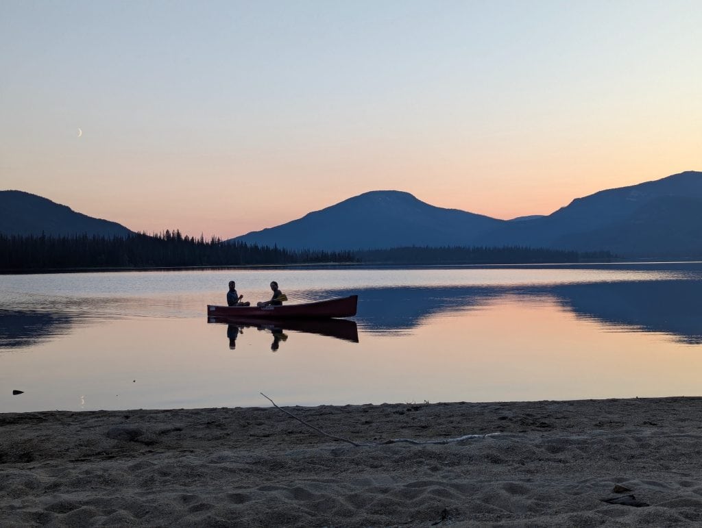 Two people are seen in a canoe at sunset, with beach in the foreground and a crescent moon hanging toward the top left of the frame, above the mountains.