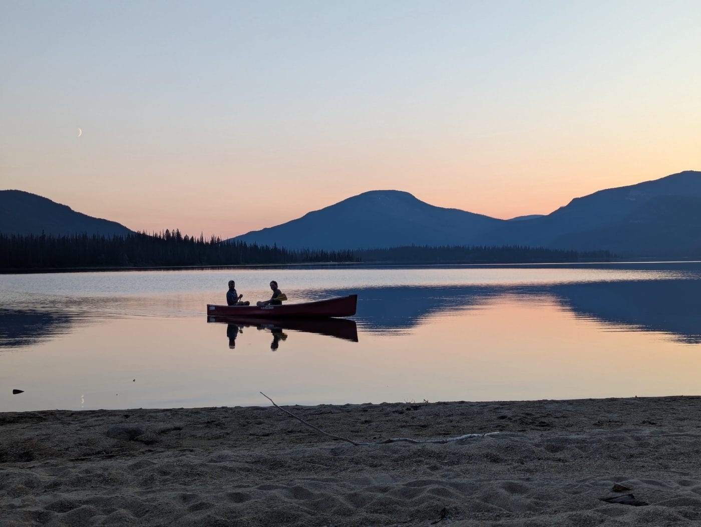 Two people are seen in a canoe at sunset, with beach in the foreground and a crescent moon hanging toward the top left of the frame, above the mountains.