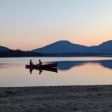 Two people are seen in a canoe at sunset, with beach in the foreground and a crescent moon hanging toward the top left of the frame, above the mountains.