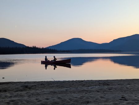 Two people are seen in a canoe at sunset, with beach in the foreground and a crescent moon hanging toward the top left of the frame, above the mountains.