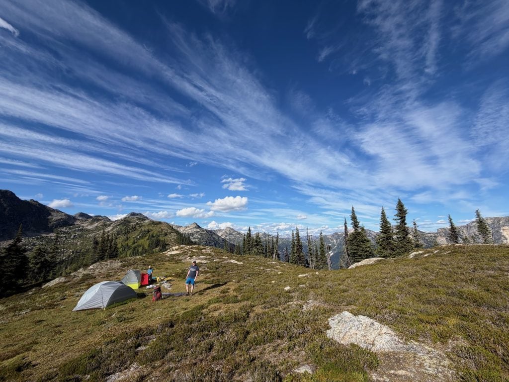 Two people are seen by their tents in a grassy patch in the alpine at Wavy Ridge in Wells Gray Provincial Park, with mountains and trees set behind.