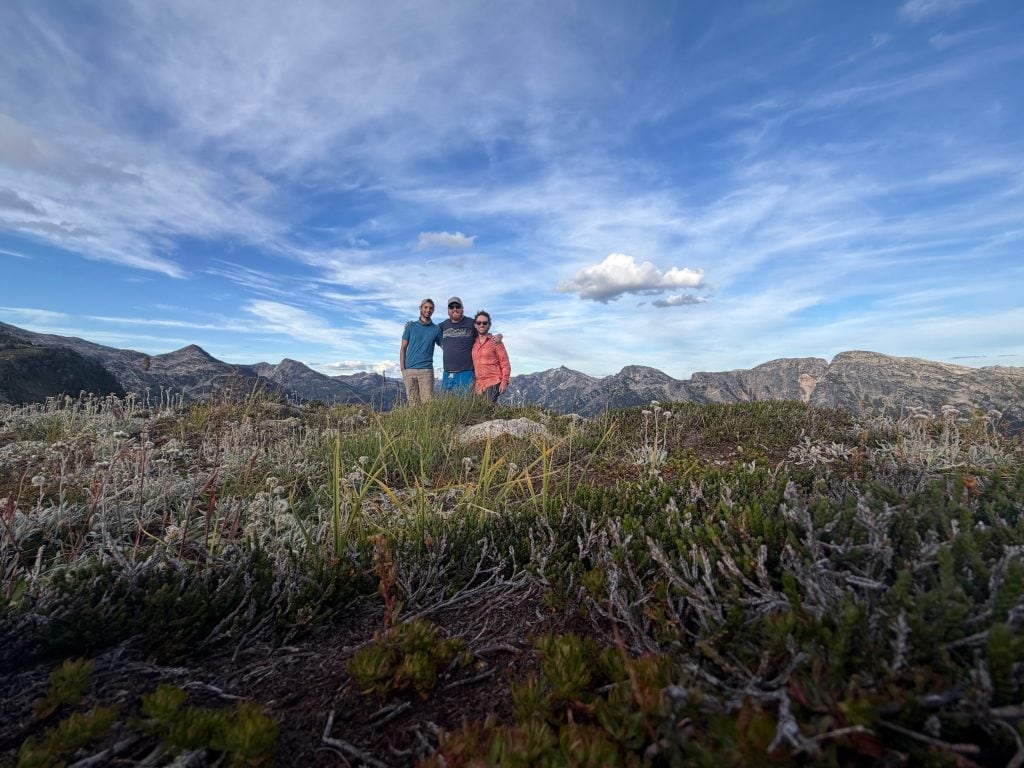 Three people pose for a photo with shrubbery in the foreground and mountains in the background, with a mix of sun and clouds.