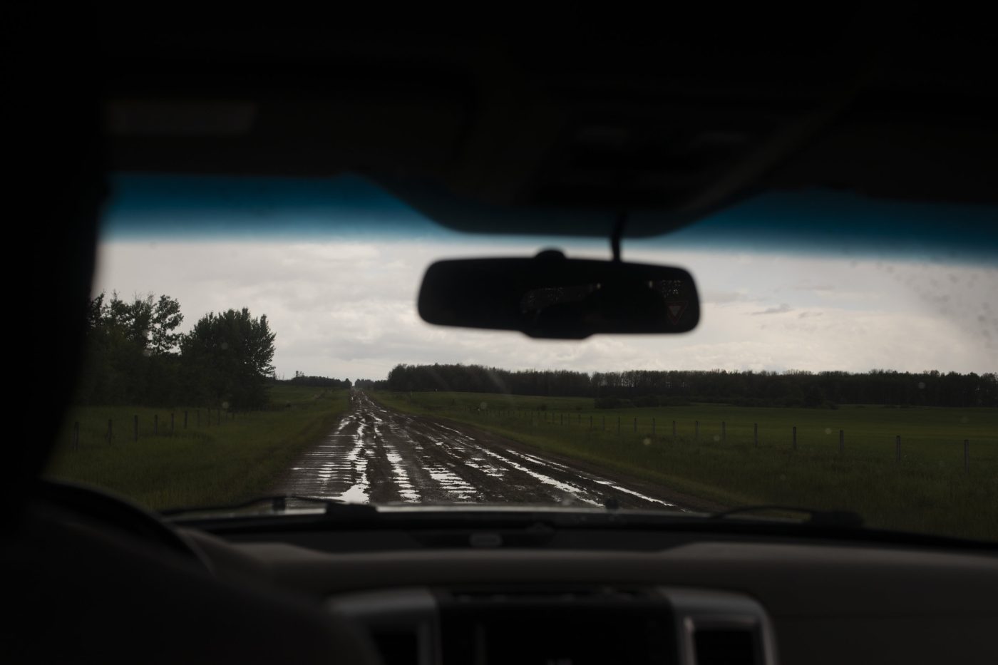 A gravel road with puddles in potholes is seen from the front seat of a car. The mood is dreary and dark.