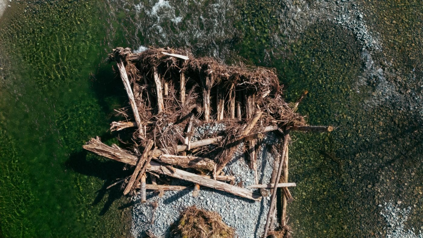Seen from above, a pile of logs and other wood debris in the middle of a relatively shallow flowing river.
