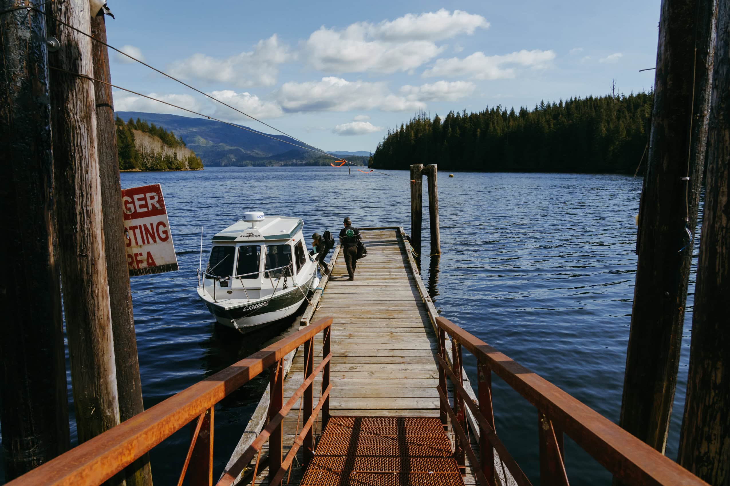 A white boat is moored beside a dock extending out into a body of water. Two people are walking on the dock.