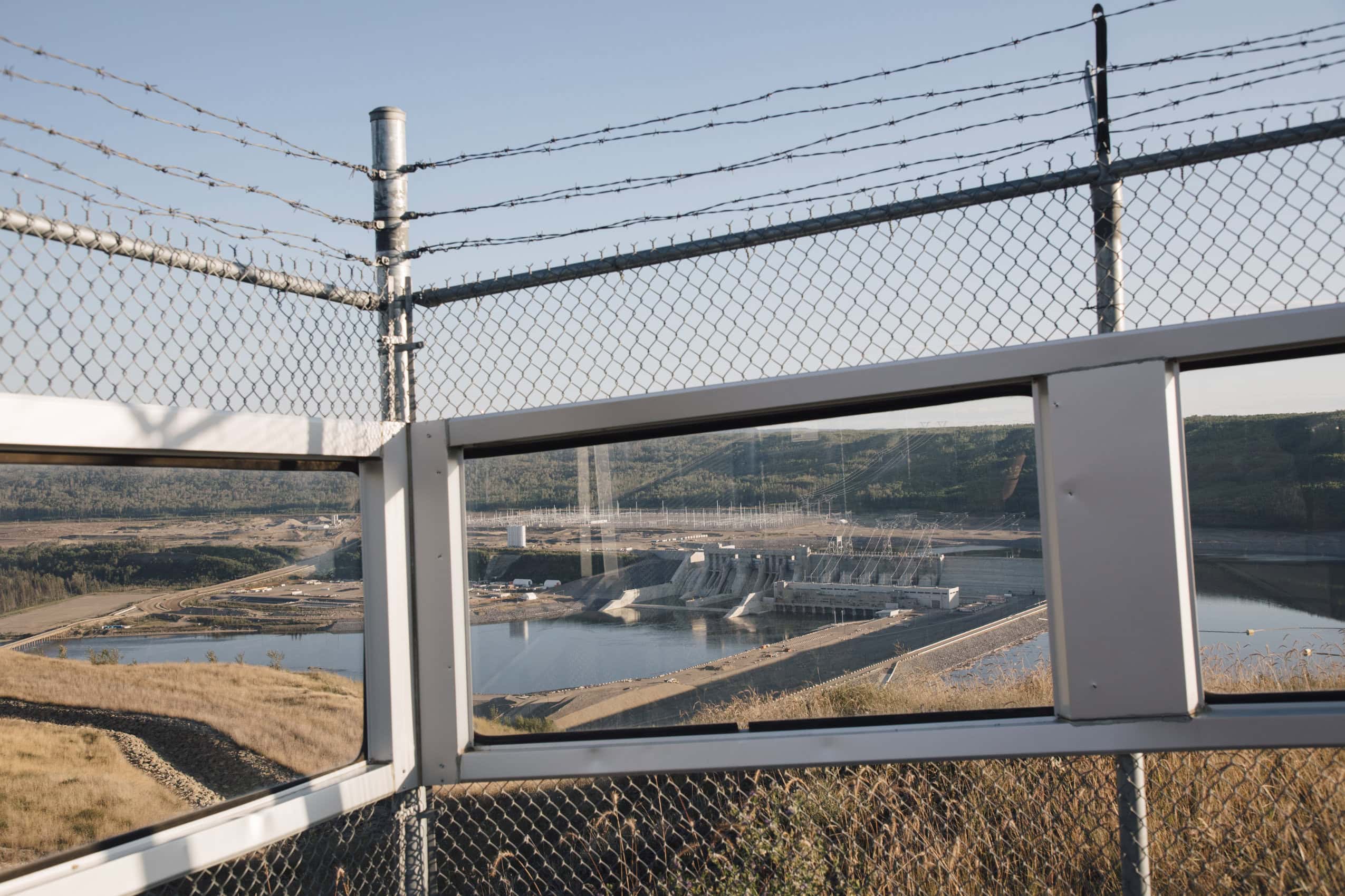 British Columbia's John Horgan dam is photographed from a lookout. The dam is framed by a window and a chain-link fence with barbed wire on top.