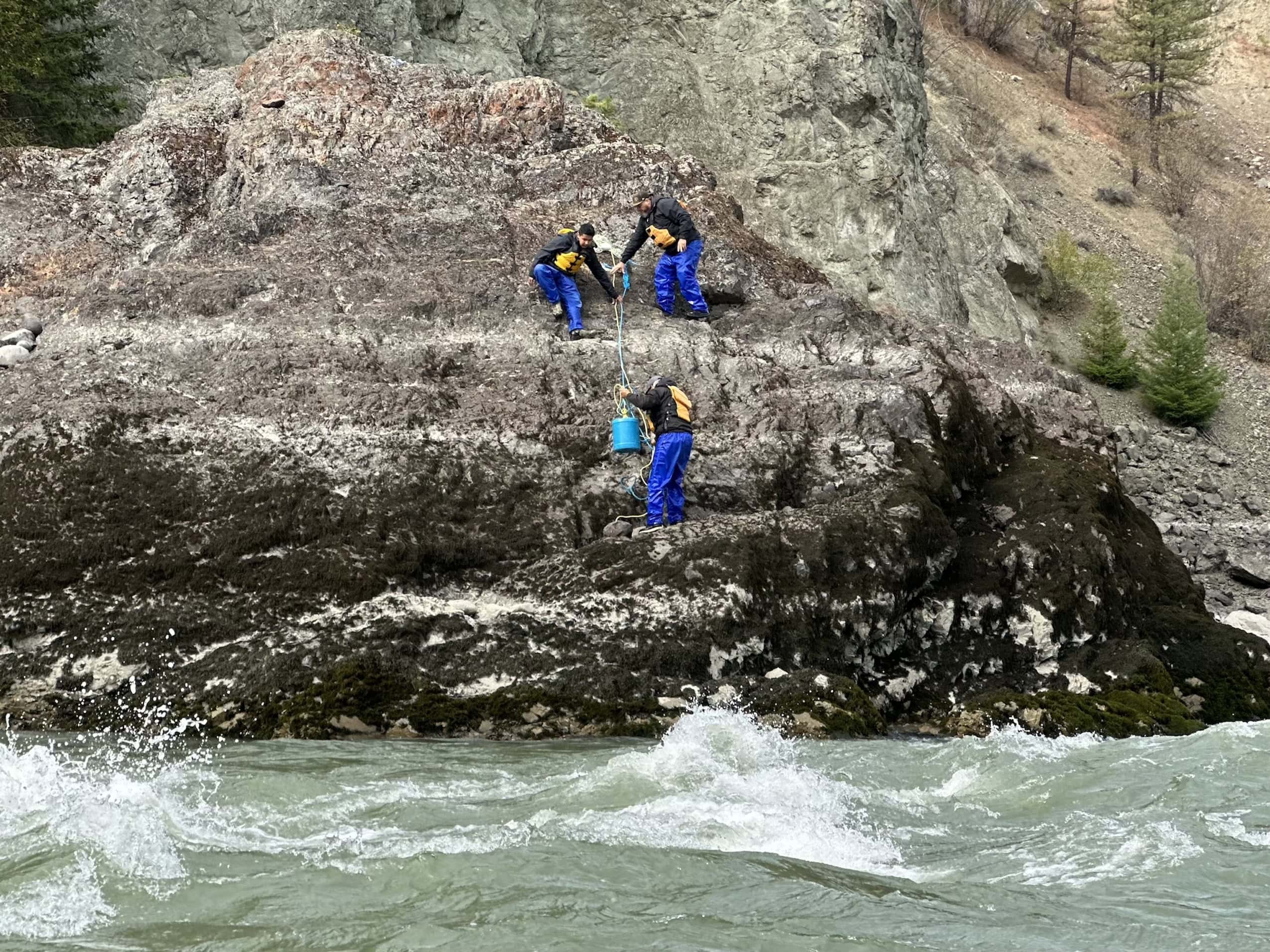 Three people wearing life jackets and rain pants scramble up the rocky shoreline of a river.