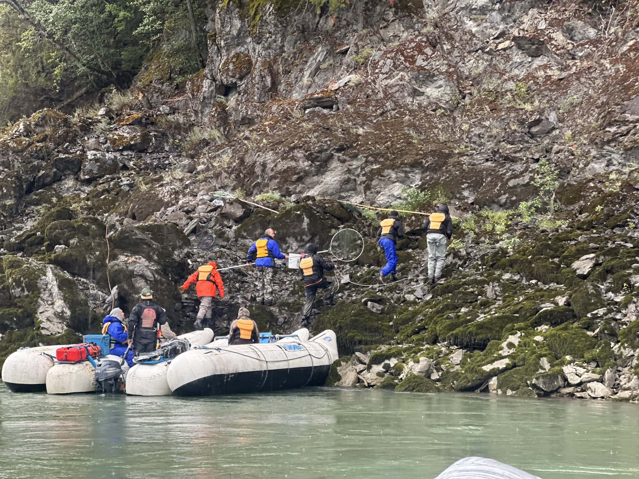 A group of people wearing life jackets and rain gear scramble up the rocky shoreline of a river.