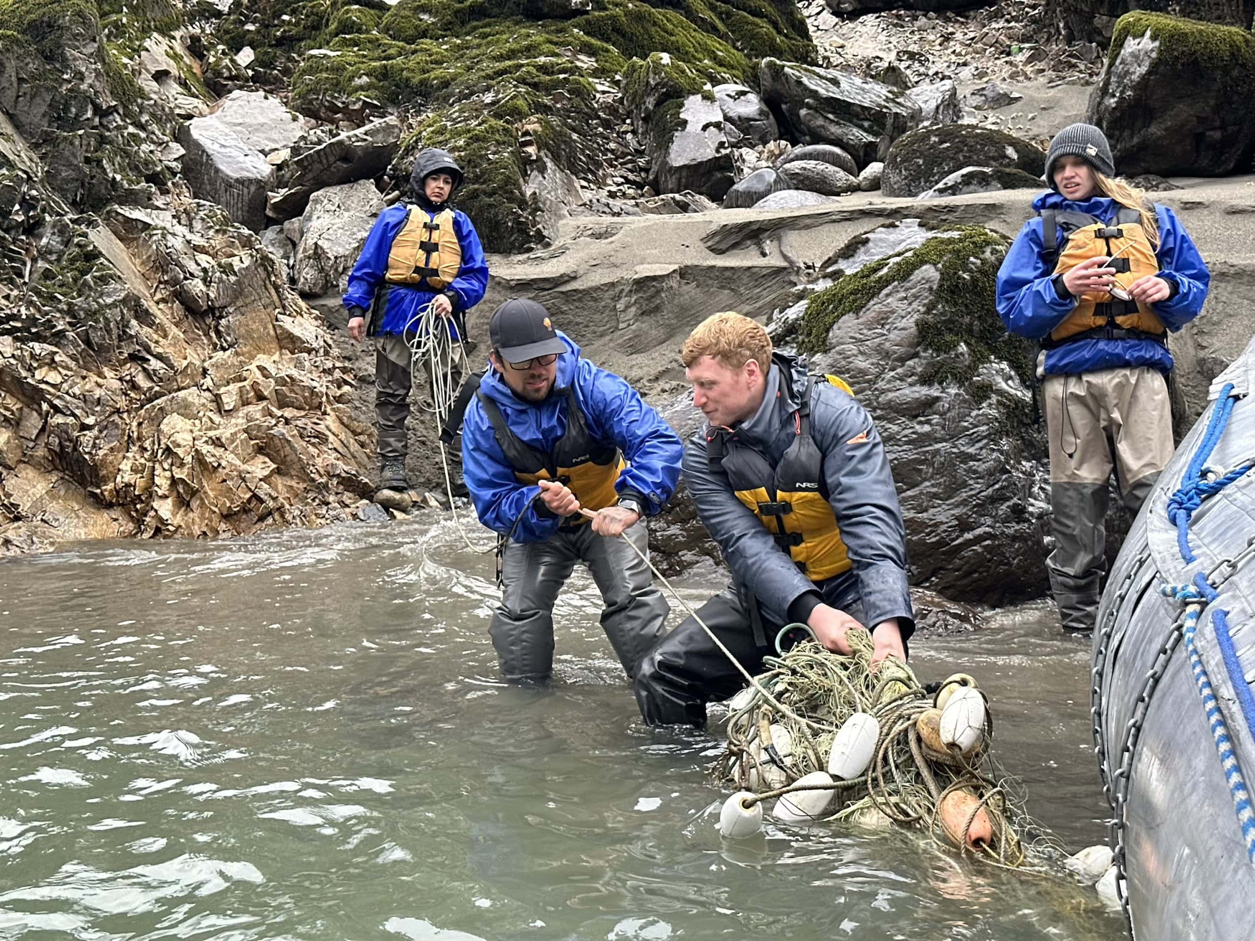 Two people stand knee-deep in a river and pull a fishing net toward them, while two others stand on shore and watch.