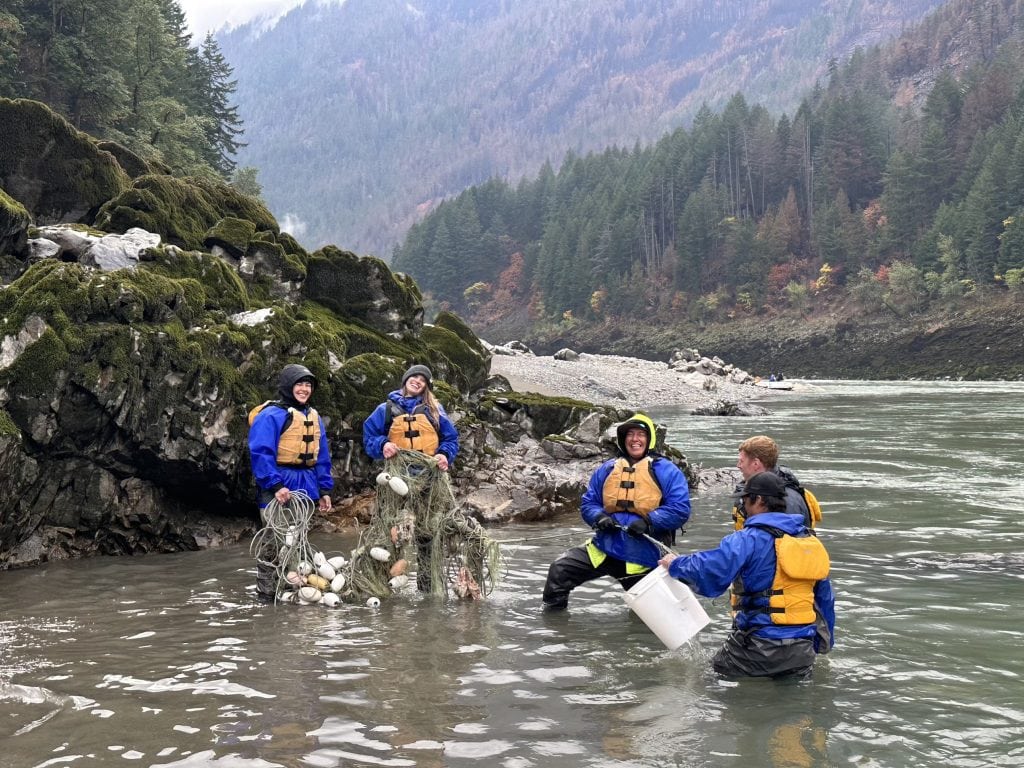 A small group of people wearing life jackets and rain gear stand in a river, smiling and holding old fishing nets.