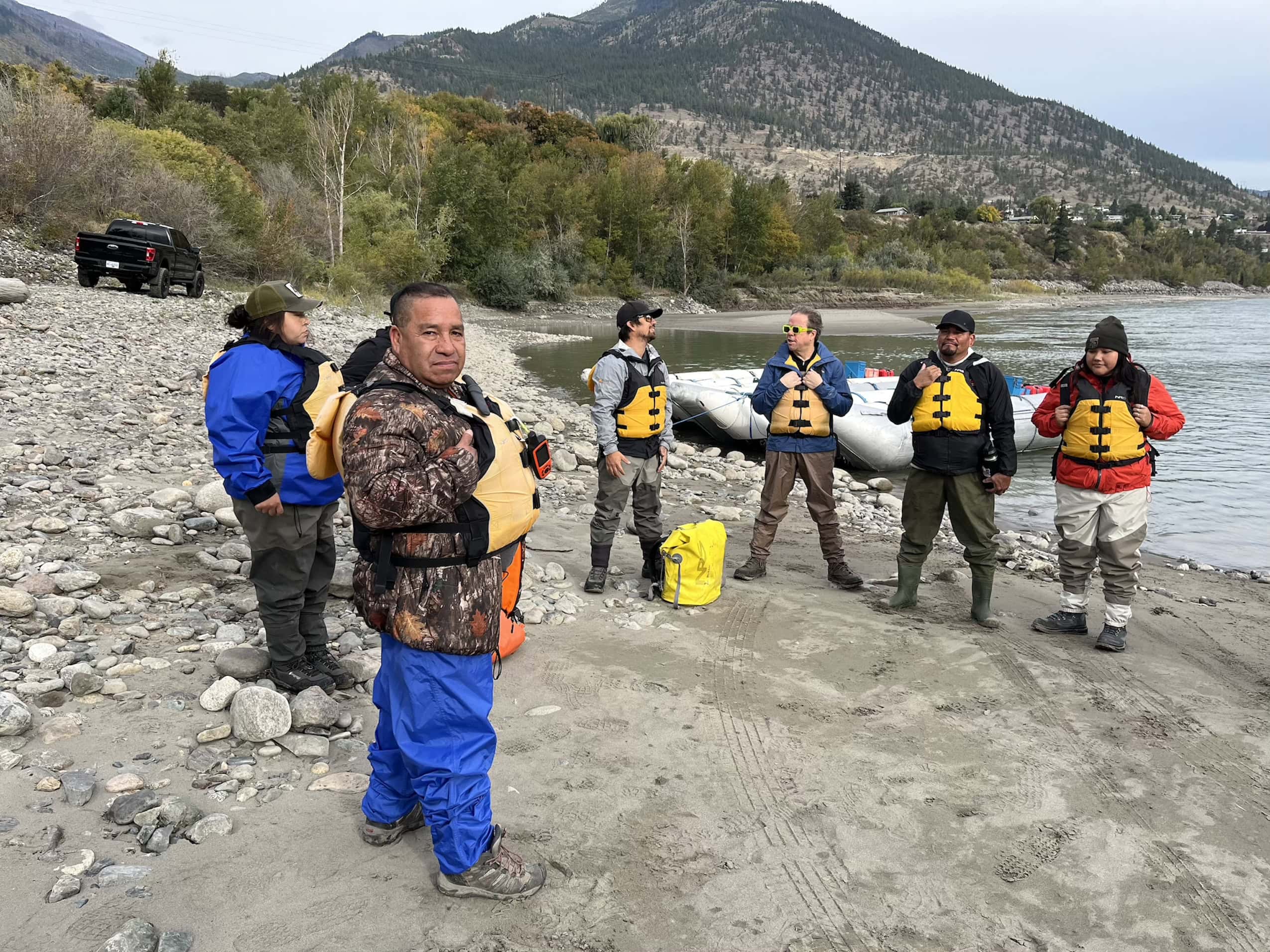 Six people stand by the shore of a river wearing life jackets.