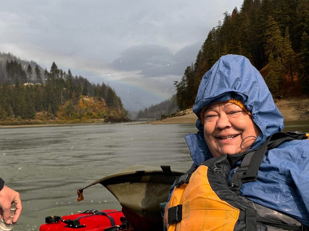 A woman wearing a hooded raincoat and a life jacket smiles, with a river in the background.
