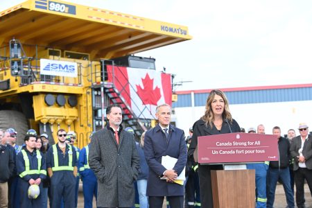 A woman (Alberta Premier Danielle Smith) speaks at a lectern while two men (Prime Minister Mark Carney and Pembina Pipeline Corp CEO Scott Burrows) stand behind her. Behind all of them is a line up of men in construction gear and casual ware. Behind all of them is a big, yellow drive mining truck.