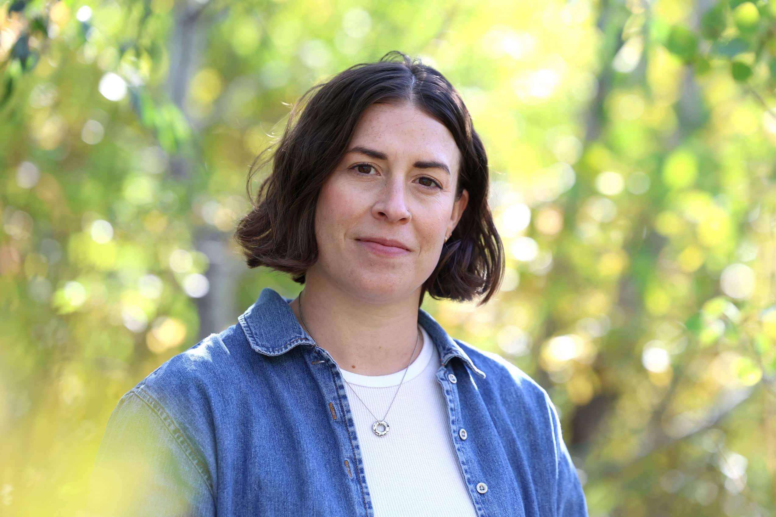 a portrait of a woman in a blue button-up and a white tee shirt standing amongst green trees.