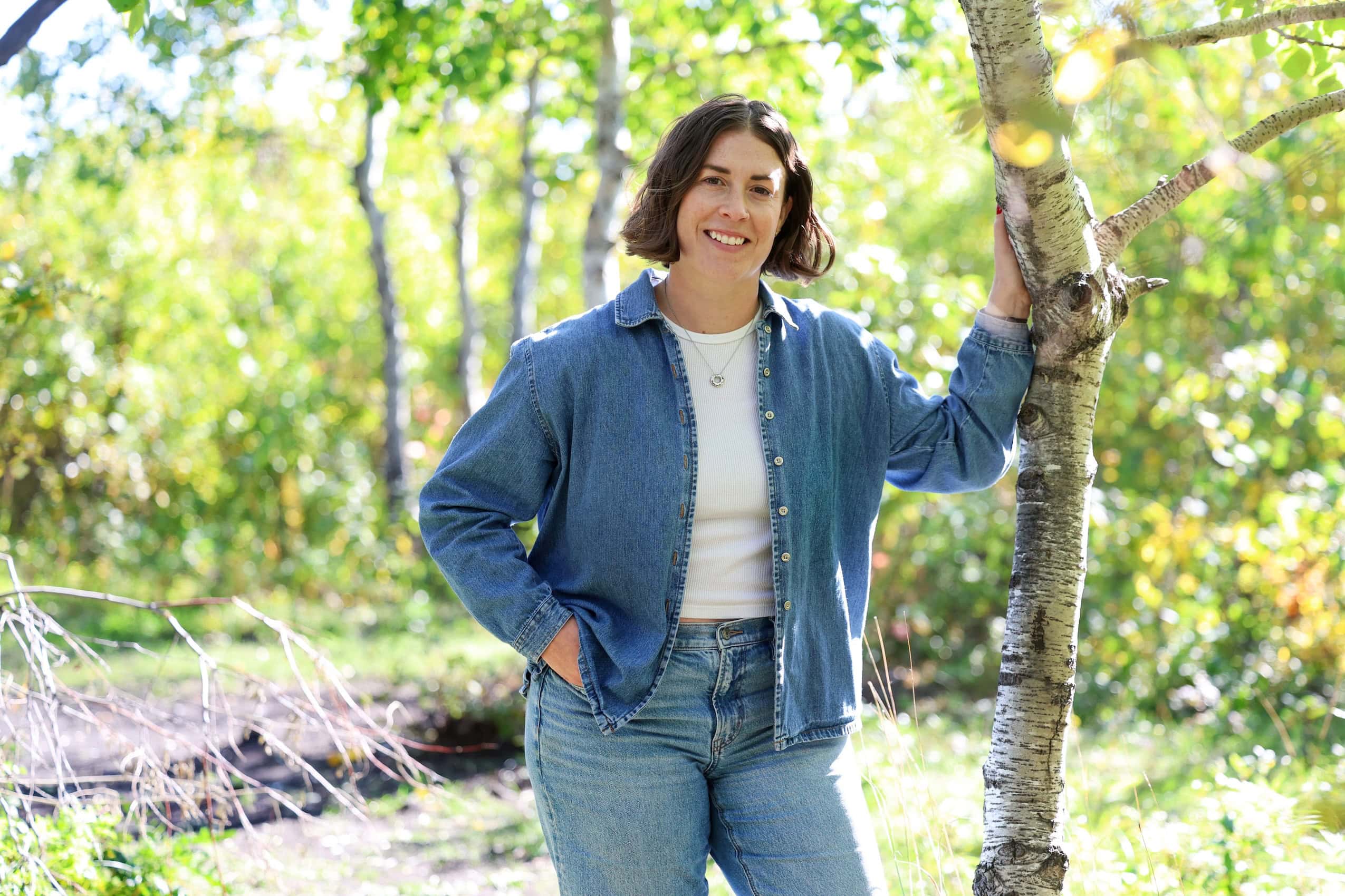 A portrait of a woman standing amongst green trees wearing a blue button-up and a white tee shirt.