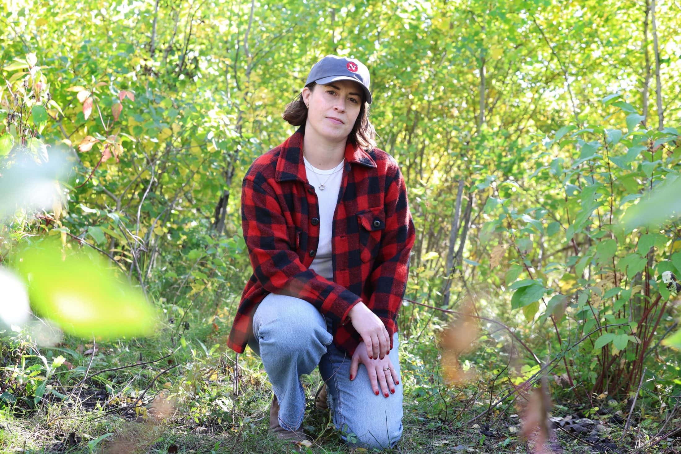 A portrait of a woman kneeling amongst green trees wearing a grey baseball cap with a red Narwhal logo, a buffalo check flannel, a white tee shirt and light blue jeans.