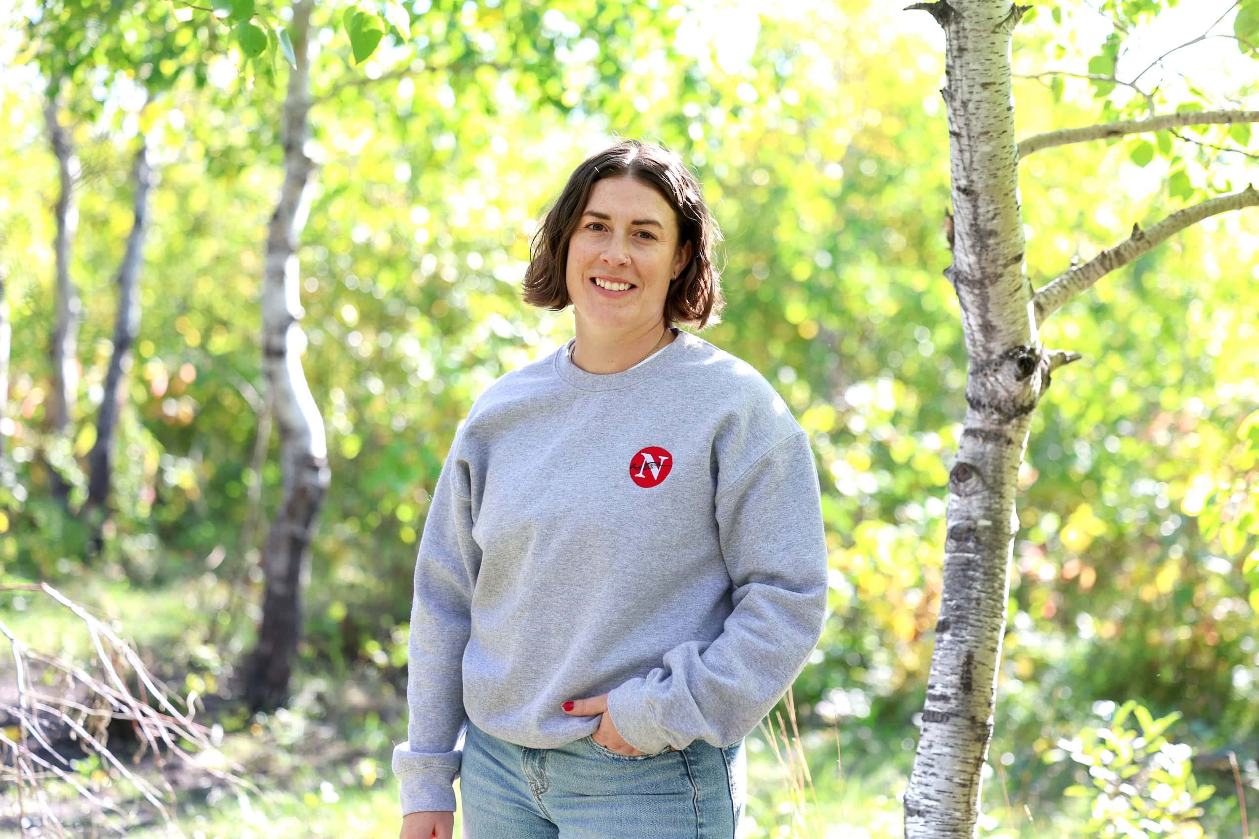 A portrait of a woman standing amongst green trees wearing a grey sweatshirt with a red Narwhal logo and light blue jeans.