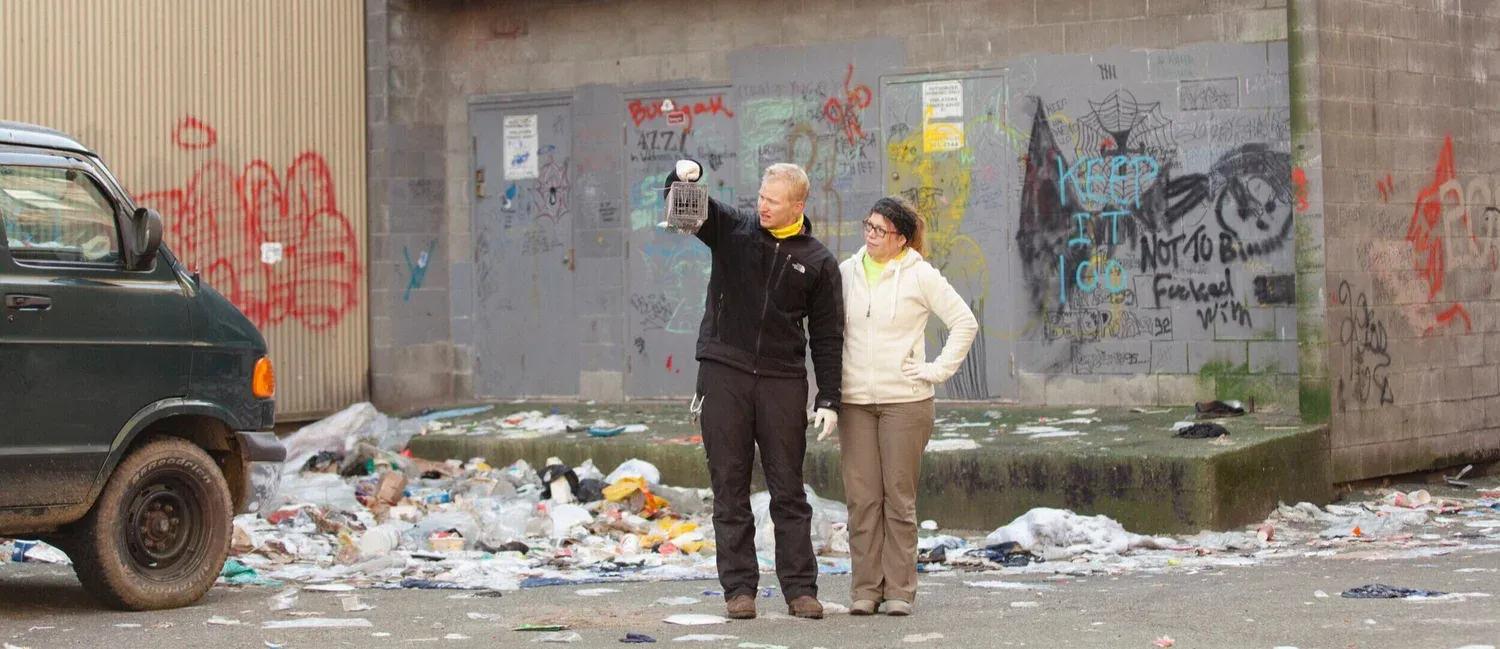 Two people stand surrounded by garbage holding a rat in a cage