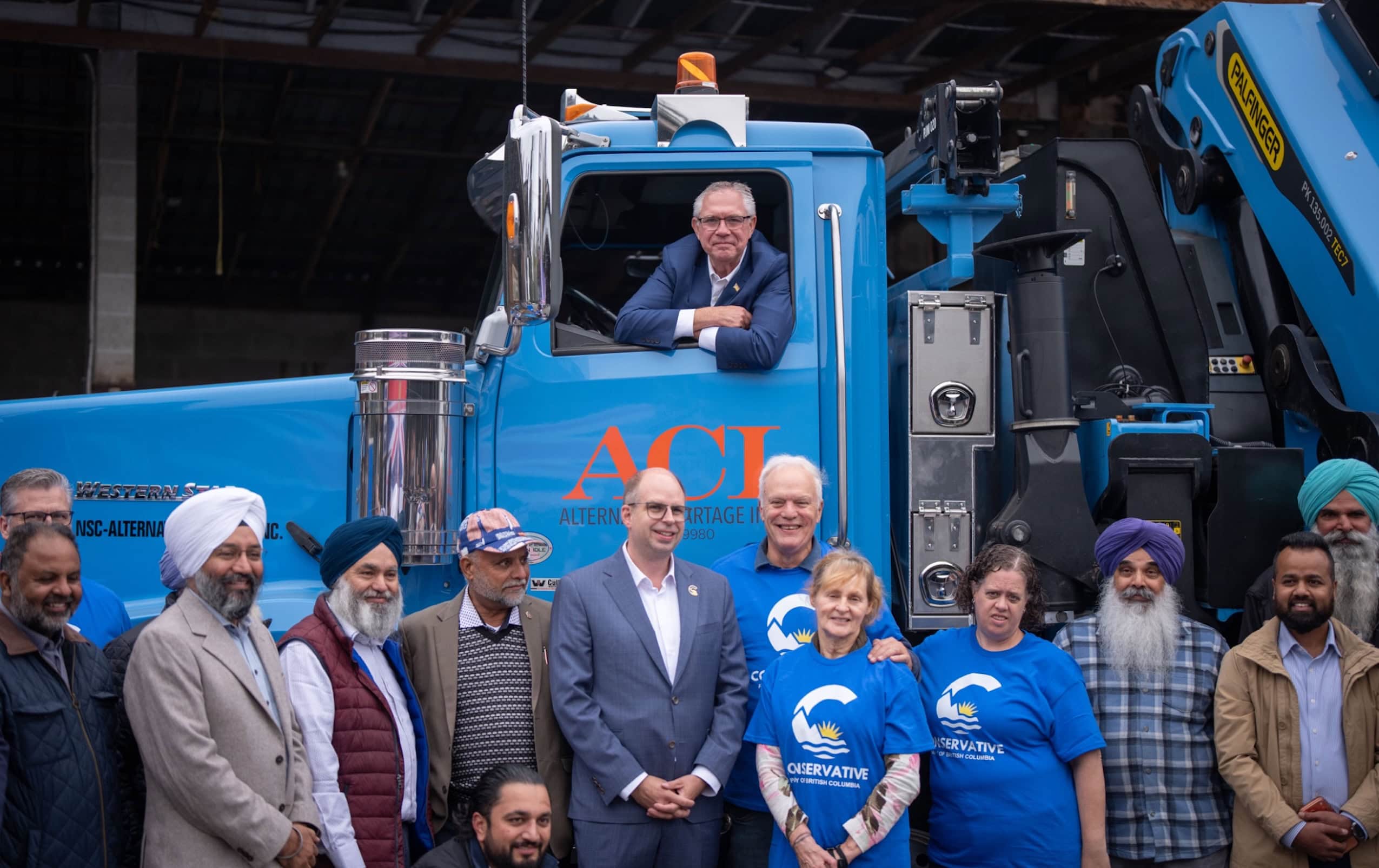 A man in a blue big rig truck looks at the camera while a line of people stand below him.