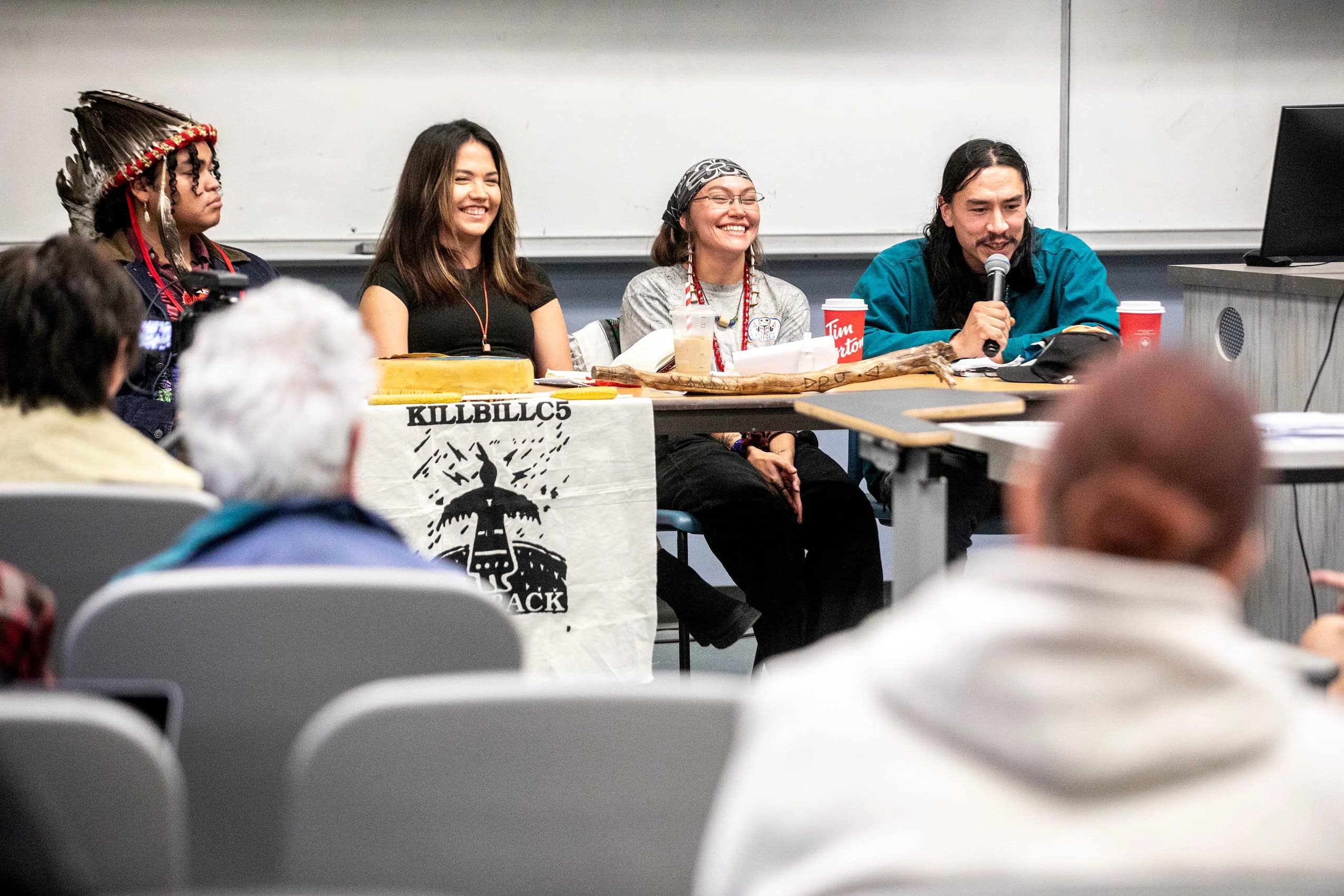 Four people sit at a table at the front of a university classroom during a panel discussion. One of them is holding a microphone.
