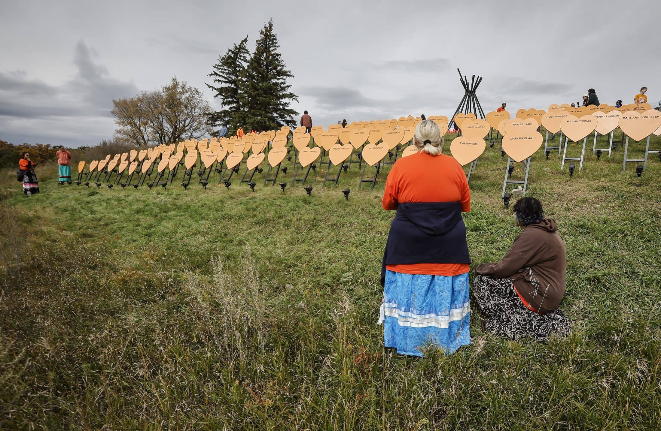 People pay respects at a memorial during an Orange Shirt Day event at the Brandon Residential School site in Brandon, Man., in 2026. They are standing in a field where rows of orange hearts bear the names of children who died.