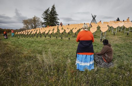 People pay respects at a memorial during an Orange Shirt Day event at the Brandon Residential School site in Brandon, Man., in 2026. They are standing in a field where rows of orange hearts bear the names of children who died.