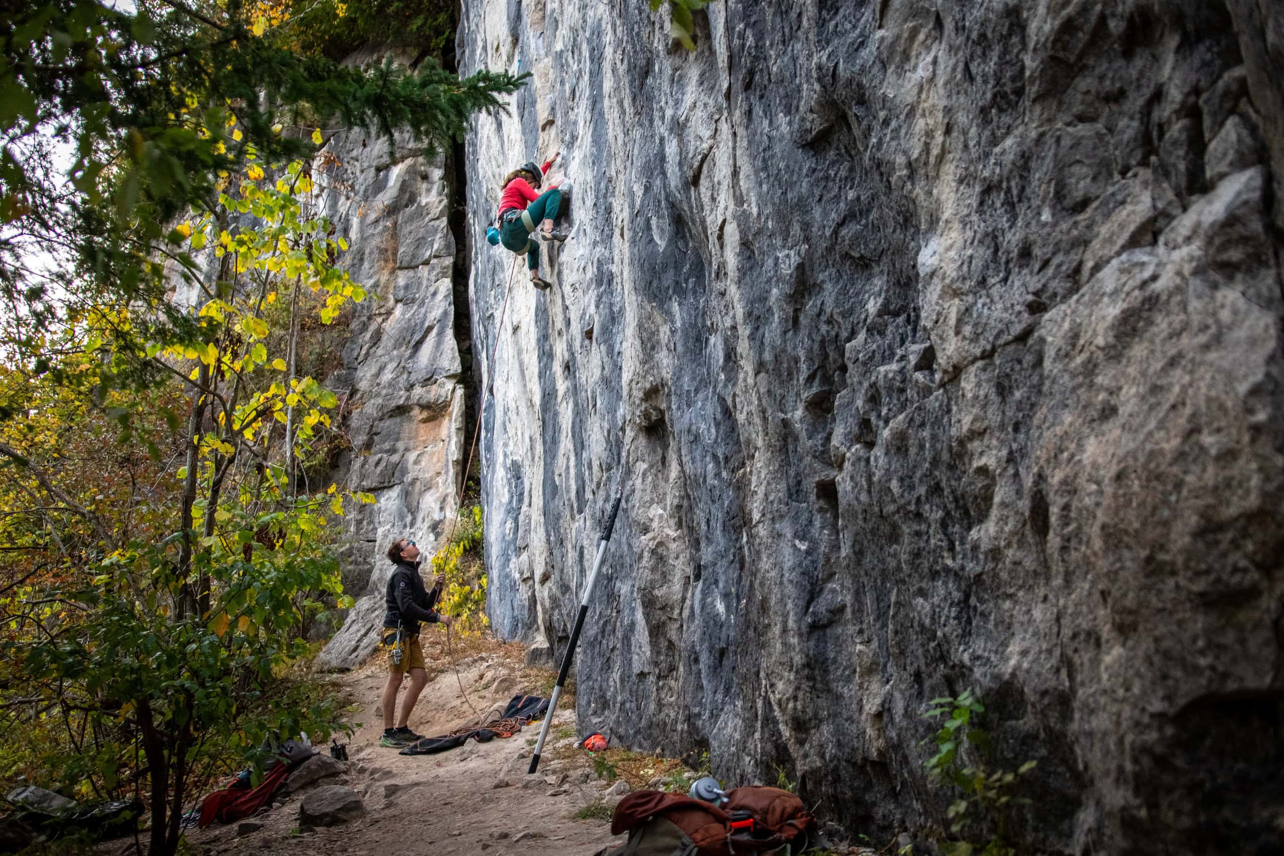 A rock climber scales a nearly-vertical rock wall, while a spotter watches from below.