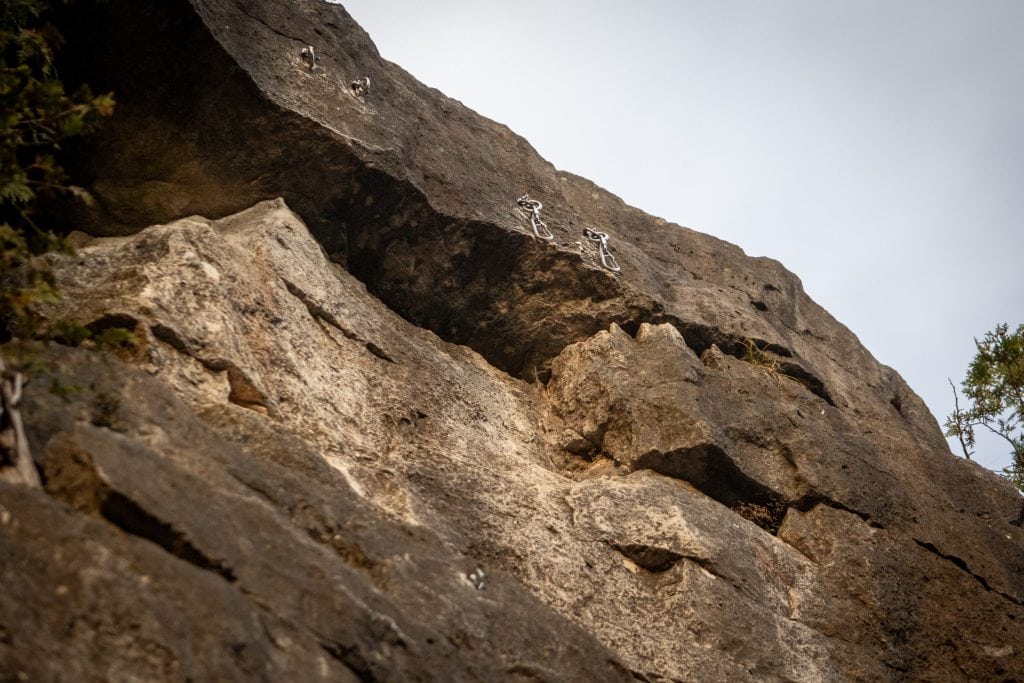 A closeup of anchors affixed near the top of a steep and rocky cliff, with climbers' carabiners hanging from them.