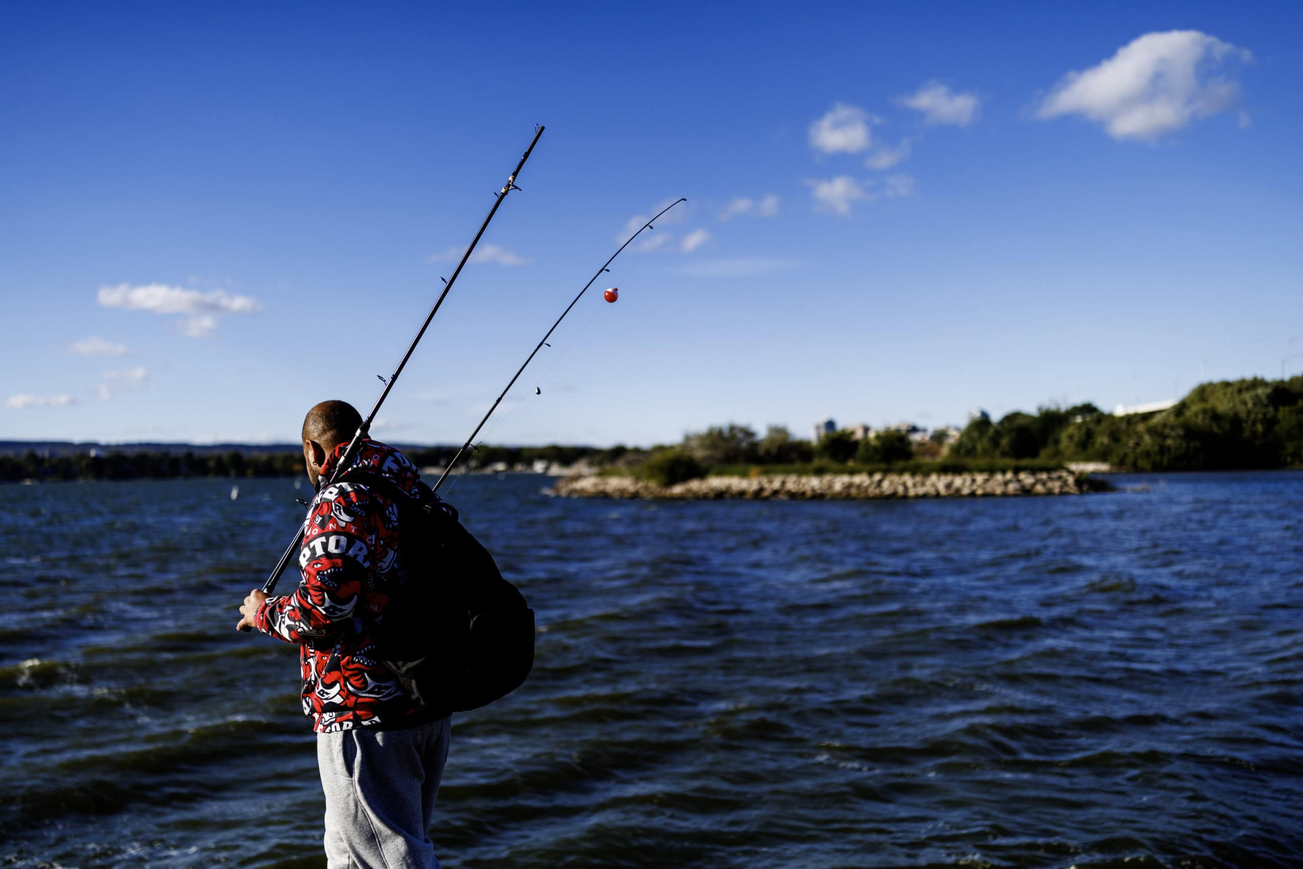 Seen from behind, a person carries fishing rods along the shore of Hamilton Harbour in Burlington, Ontario.