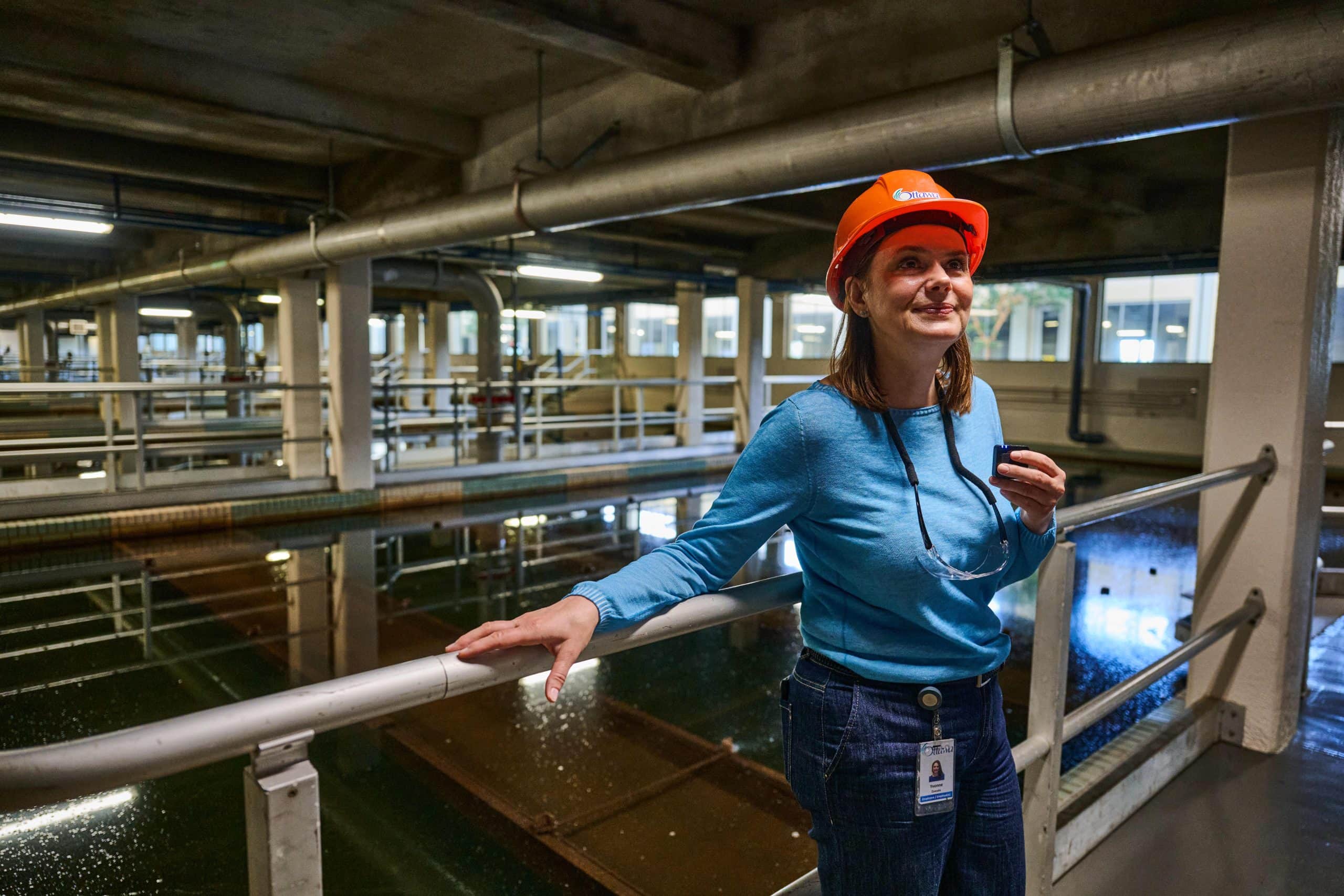 Woman in blue shirt and orange safety helmet smiles and leans against a railing with large water tanks in the background.
