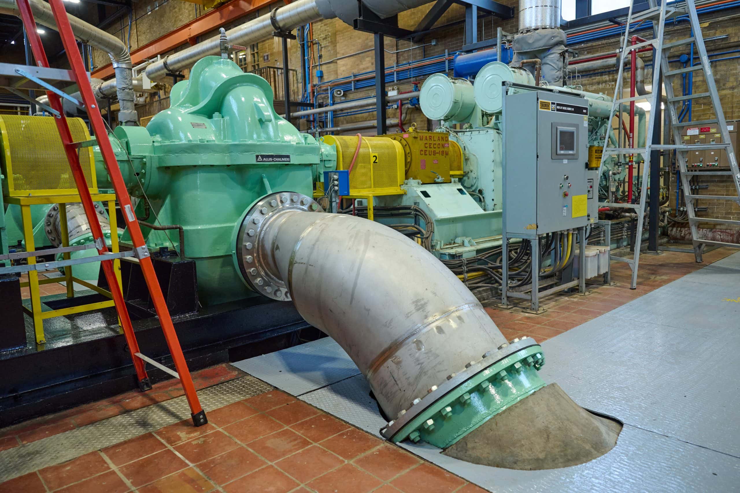 Large green and grey industrial pump inside a warehouse attached to a pipe that goes into the floor on an angle.
