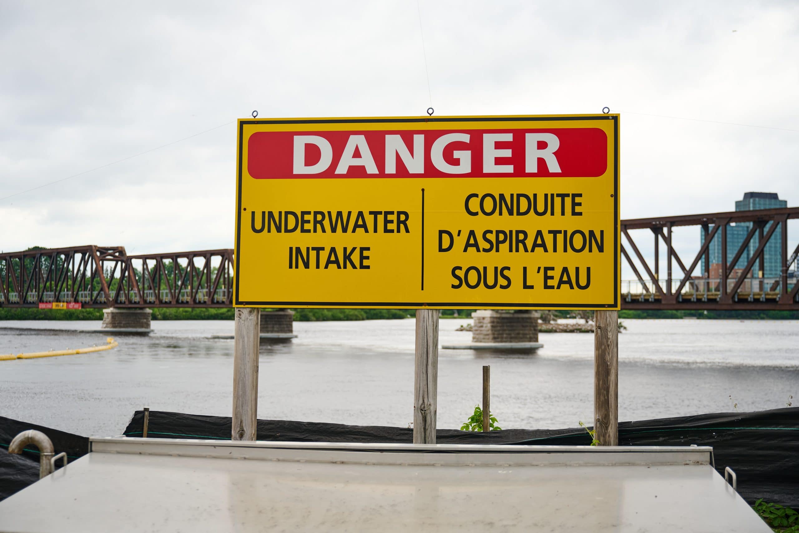 Big yellow and red sign that says "Danger: underwater intake" with a bridge in the background crossing over a river.